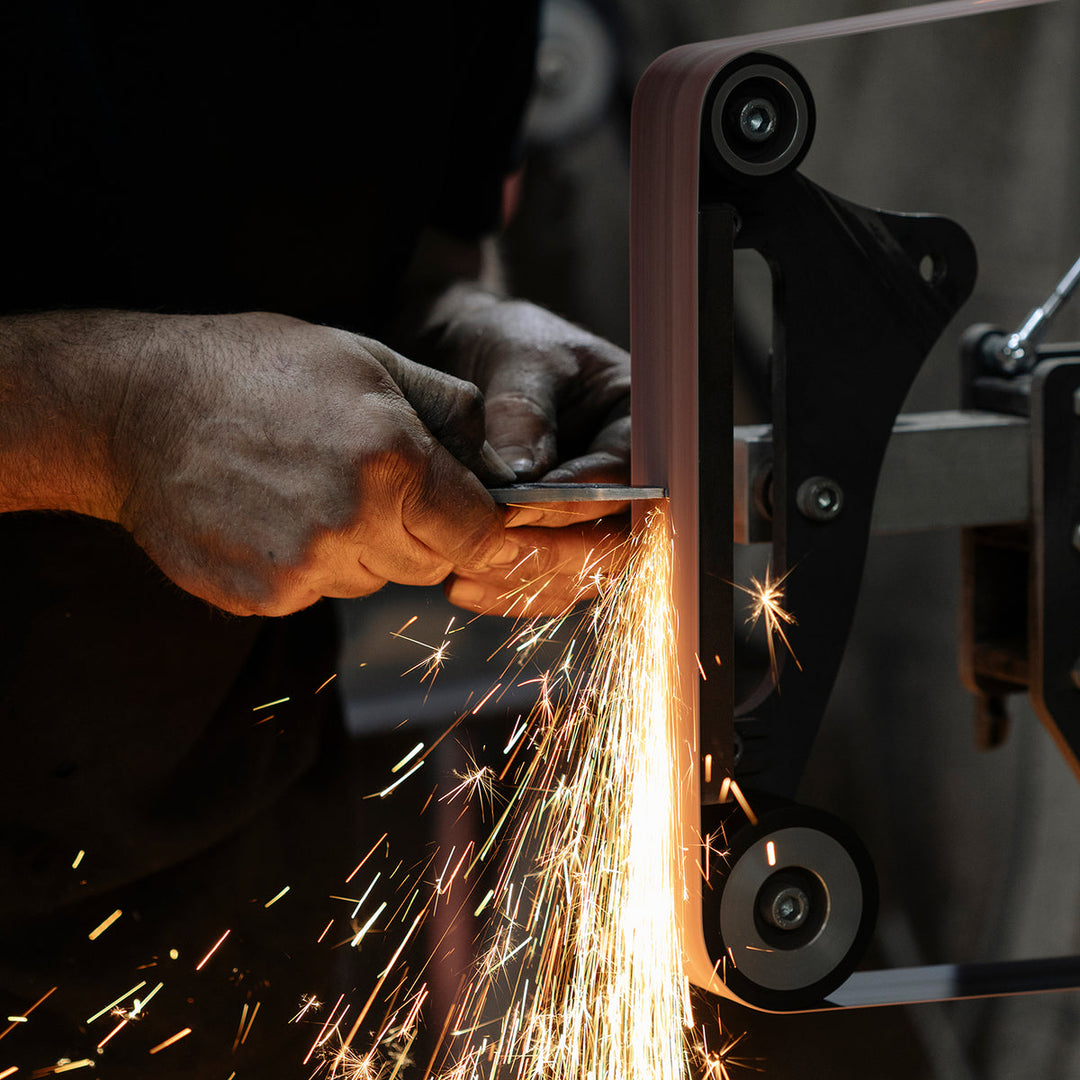 A close-up action shot of a person using a belt sander to sharpen a metal file, with bright orange sparks flying. The person's hands and arms are visible, and the machine is shown in use against a dark, industrial background.