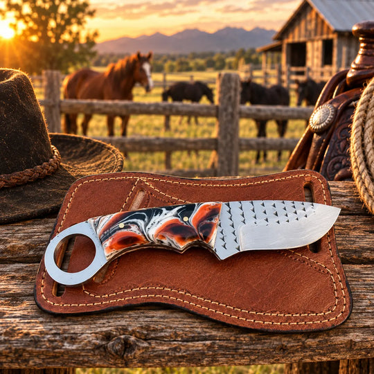 A fixed-blade knife with a marbled black, orange, and white handle and a textured silver blade rests in a brown leather sheath on a rustic wooden table. A cowboy hat is partially visible on the left, and a saddlery setup with rope and a horse is blurred in the background at sunset.