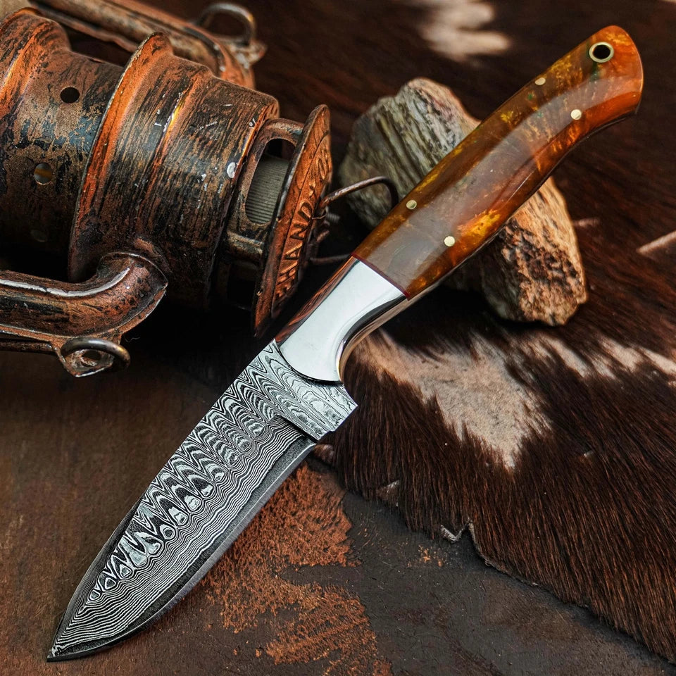 A fixed-blade knife with a patterned Damascus steel blade and a polished amber-brown wooden handle, displayed diagonally on a rustic background with a vintage fishing reel and textured hide.