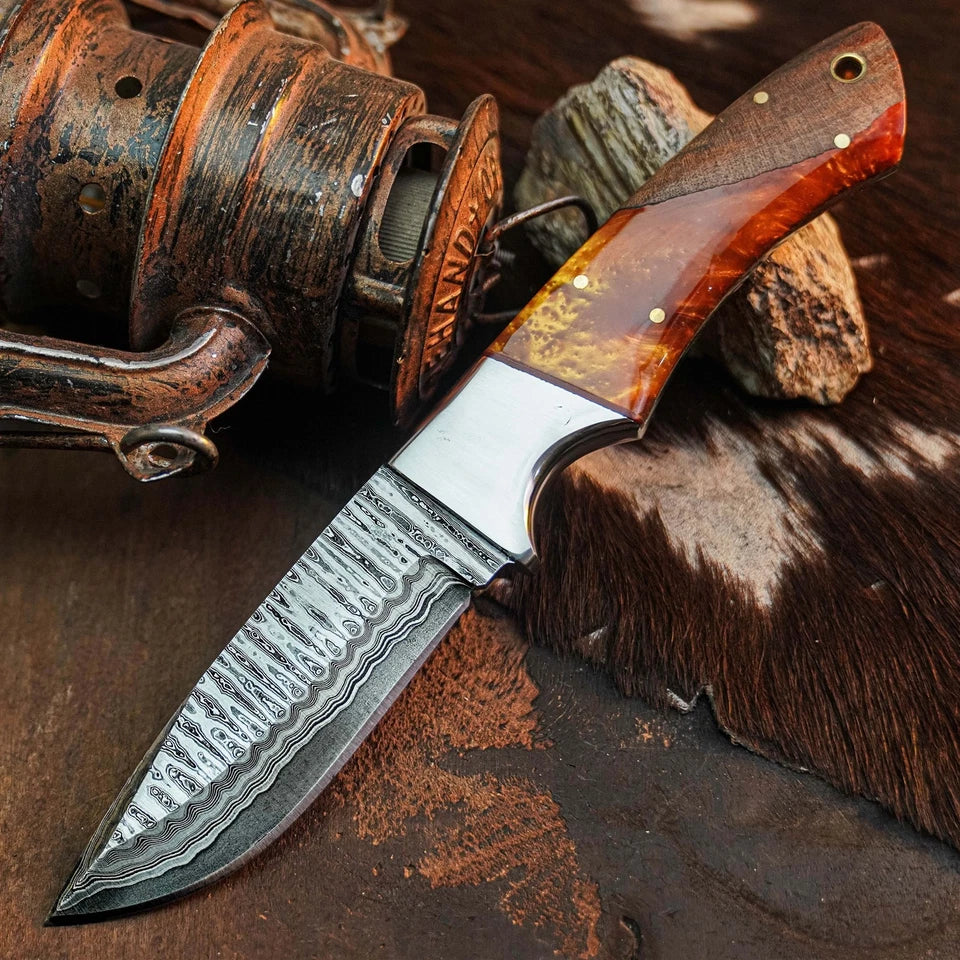 A fixed-blade knife with a patterned Damascus steel blade and a reddish-brown wood handle, displayed diagonally on a rustic dark surface next to an antique-style reel and cowhide.