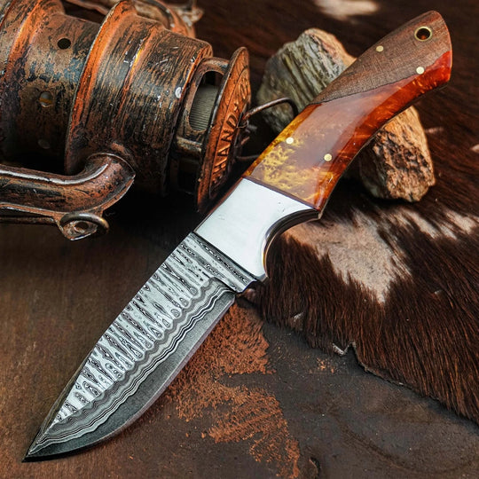 A fixed-blade knife with a patterned Damascus steel blade and a reddish-brown wood handle, displayed diagonally on a rustic dark surface next to an antique-style reel and cowhide.