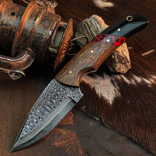 A custom Damascus steel folding knife with a patterned steel blade, a brown wood handle, and a black handle with red and gold accents, displayed on a rustic background with a vintage fishing reel and cowhide.