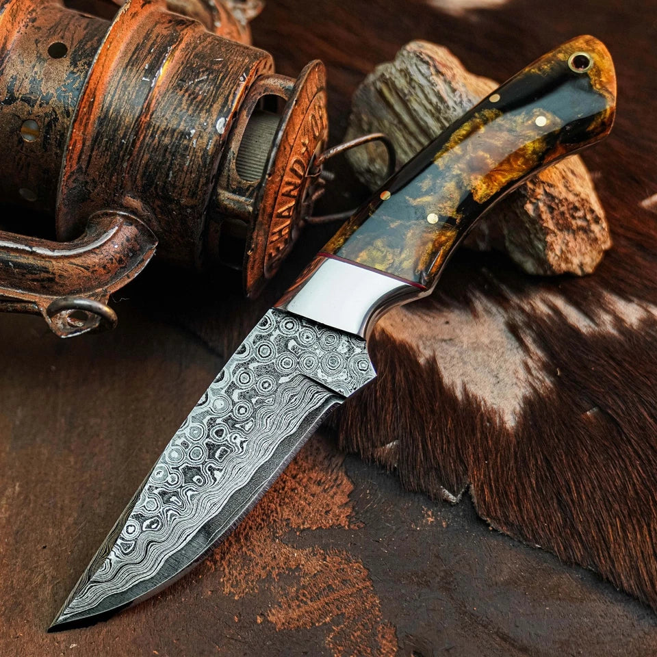 A custom Damascus steel fixed-blade knife with a patterned blade and a polished handle, displayed diagonally on a rustic background with a vintage fishing reel and cowhide.