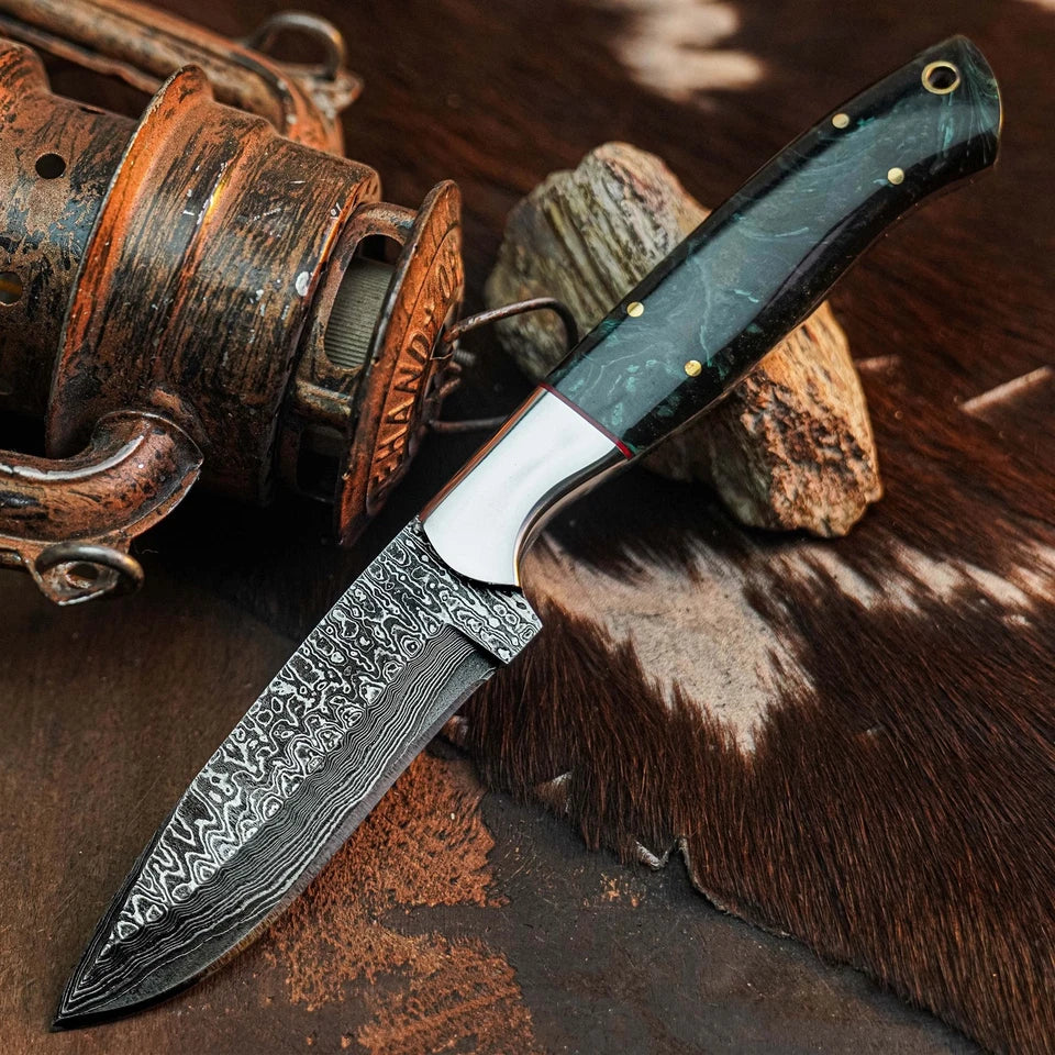 A fixed-blade knife with a patterned Damascus-style steel blade and a dark green marbled handle, displayed diagonally on a rustic brown fur surface next to a vintage wooden reel.