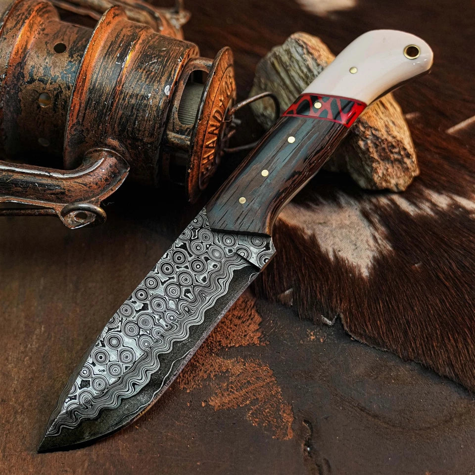 A custom fixed-blade knife with a patterned Damascus steel blade, a dark wood handle with brass pins, and a cream-colored end cap, displayed diagonally on a rustic brown leather and wood background.