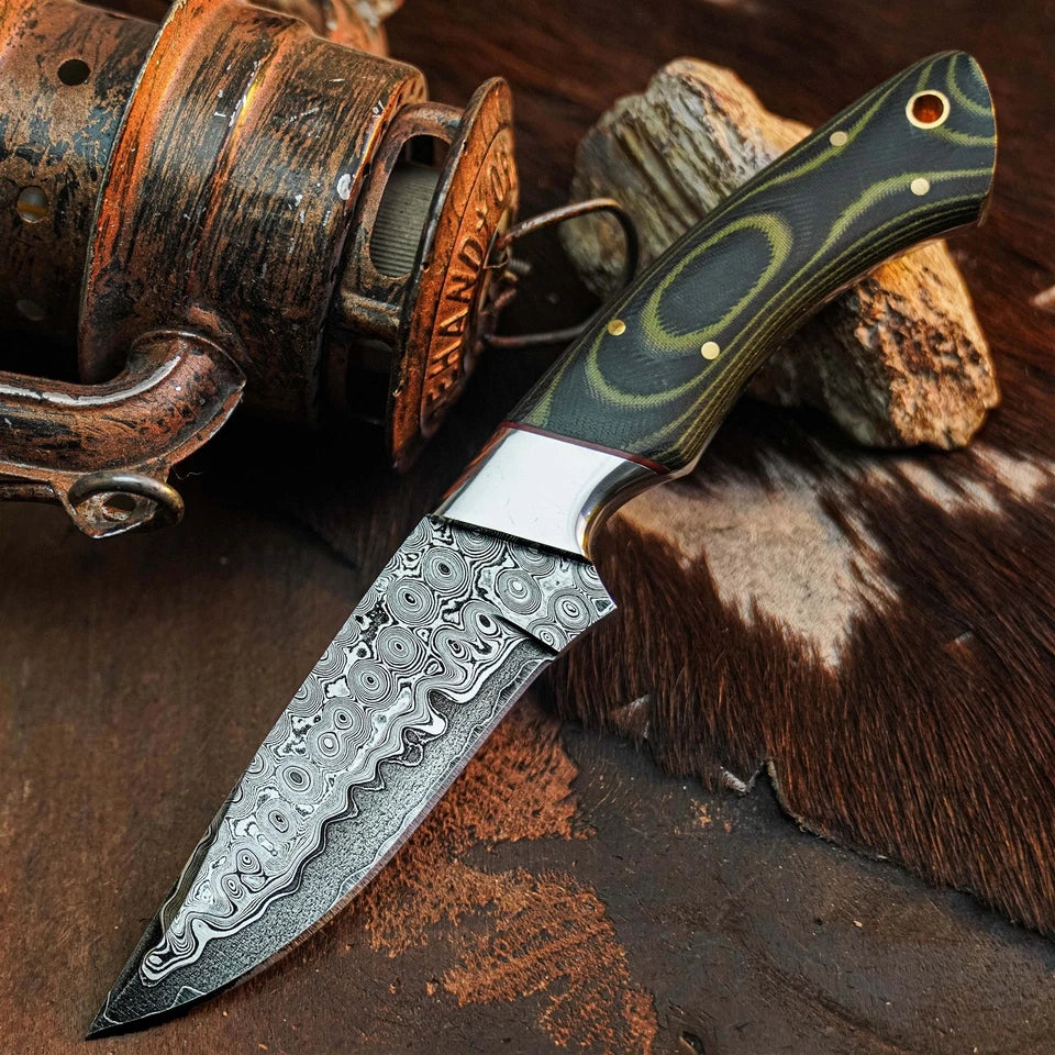 A custom Damascus steel pocket knife with a patterned steel blade and a green marbled handle, displayed on a rustic background with a vintage reel and wood.