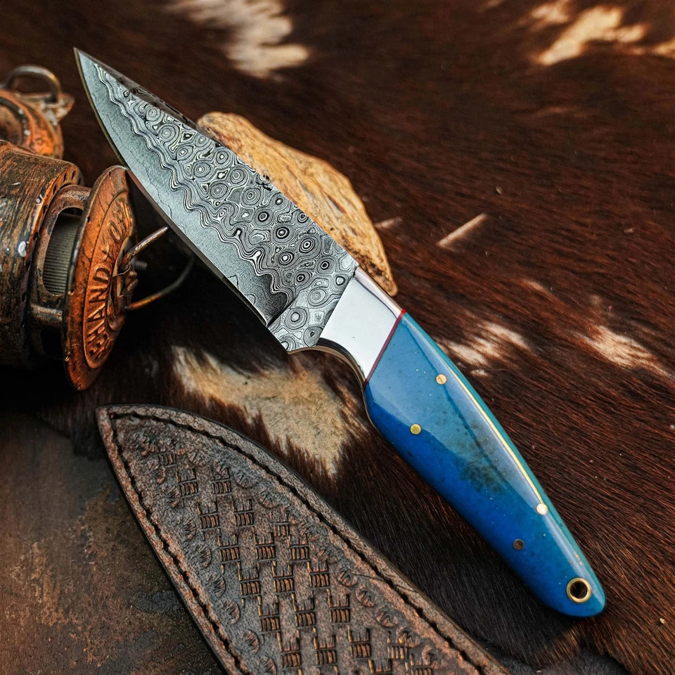 A fixed-blade knife with a patterned steel blade and a bright blue handle with brass pins, resting on a brown and white cowhide surface next to a leather sheath and a rustic metal container.