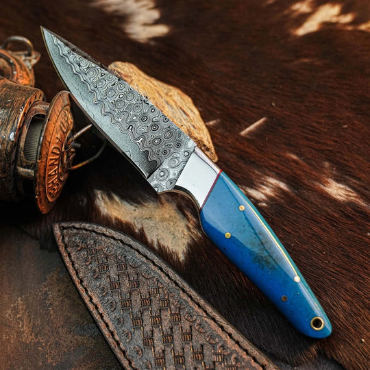 A fixed-blade knife with a patterned steel blade and a bright blue handle with brass pins, resting on a brown and white cowhide surface next to a leather sheath and a rustic metal container.