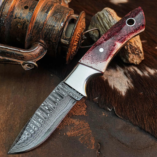 A custom fixed-blade knife with a Damascus steel blade and a reddish-brown wood handle, styled on a rustic background with a vintage fishing reel and cowhide.