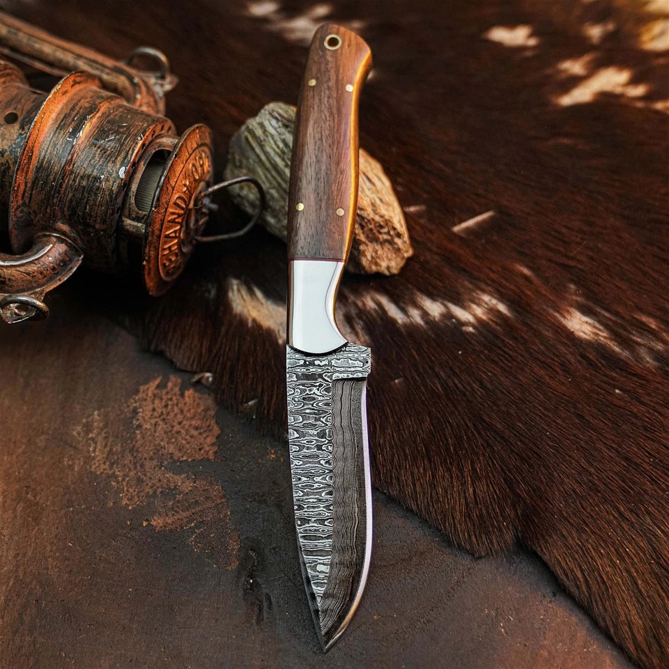 A fixed-blade knife with a patterned Damascus steel blade and a brown wooden handle, displayed on a dark textured surface next to a vintage wooden reel and a cowhide rug.