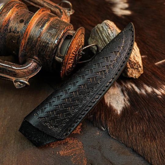 A black leather knife sheath with an embossed geometric pattern, shown next to a rustic vintage wooden reel on a textured brown and white cowhide surface.