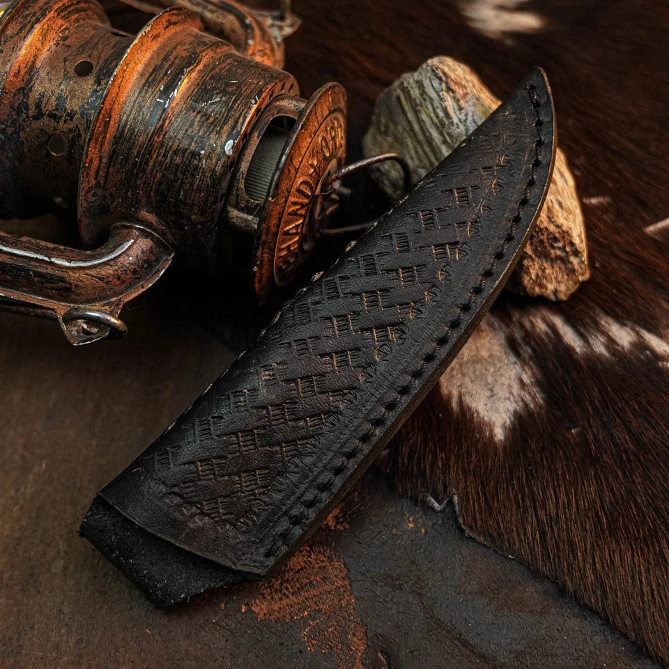 A black leather knife sheath with a woven basketweave pattern and stitched edges, shown resting on a rustic dark surface with a vintage wooden reel and cowhide accents.