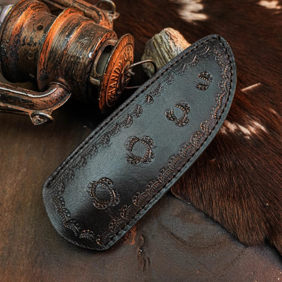 A black leather knife sheath with embossed floral and scrollwork details, displayed diagonally on a rustic brown textured surface next to an antique wooden fishing reel.