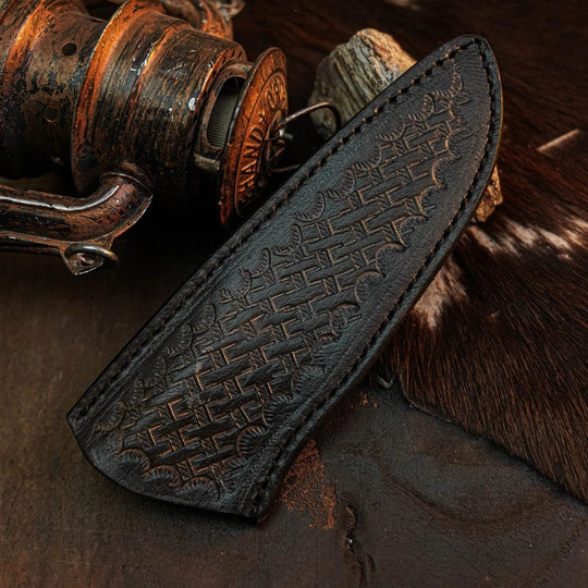 A dark brown leather knife sheath with embossed geometric patterns, shown diagonally on a rustic dark surface next to a vintage wooden fishing reel and cowhide.