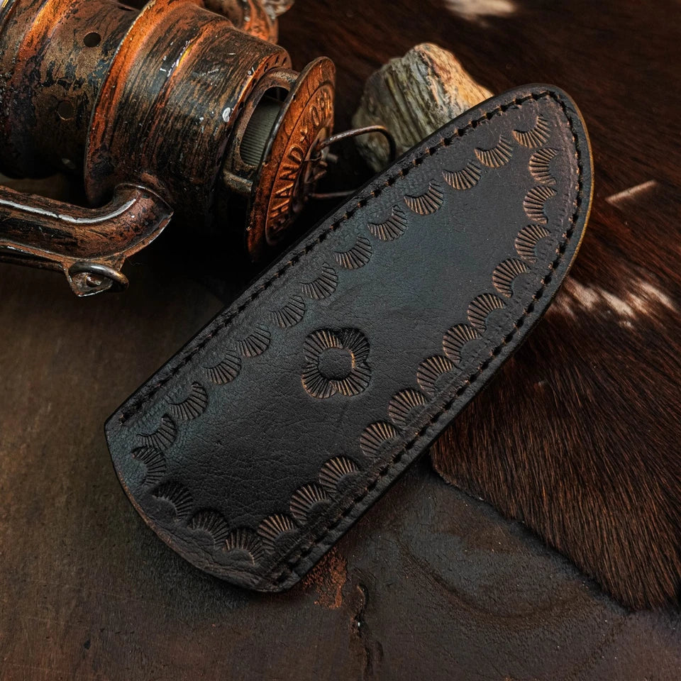 A dark brown leather knife sheath with hand-carved floral and scalloped patterns, displayed on a rustic background with a vintage wooden reel and cowhide.