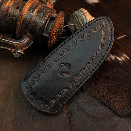 A dark brown leather knife sheath with hand-carved floral and scalloped patterns, displayed on a rustic background with a vintage wooden reel and cowhide.