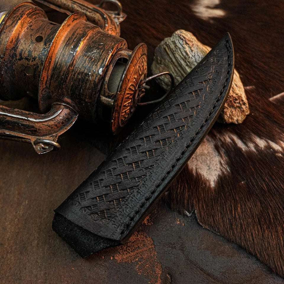 A black leather knife sheath with a textured basketweave pattern and visible stitching, shown resting on a rustic surface next to a vintage copper-toned fishing reel and cowhide.
