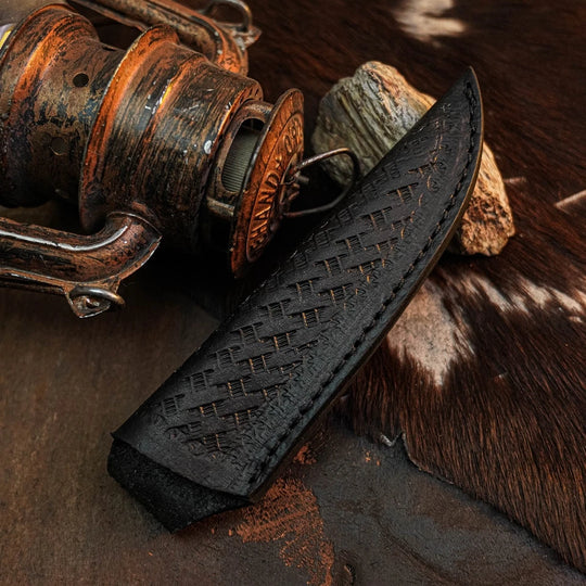 A black leather knife sheath with a textured basketweave pattern and visible stitching, shown resting on a rustic surface next to a vintage copper-toned fishing reel and cowhide.