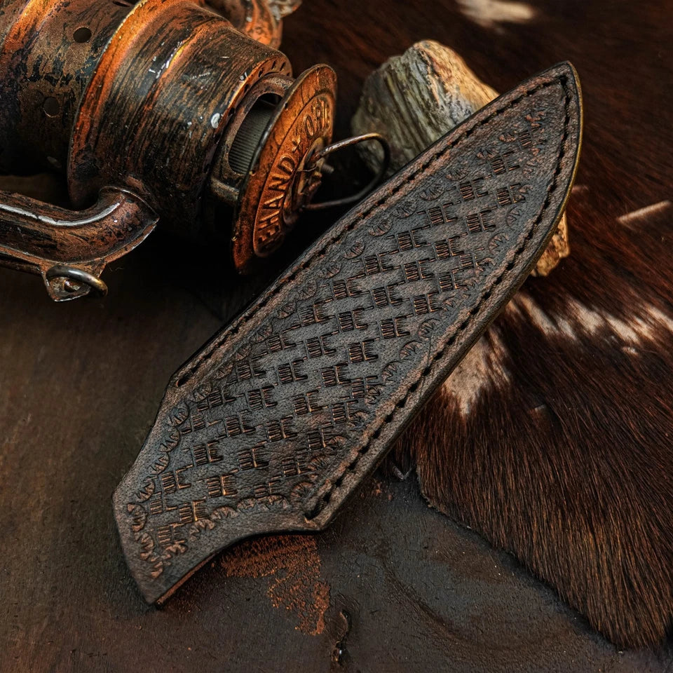 A dark brown leather knife sheath with an embossed geometric pattern, displayed diagonally on a rustic dark surface next to a vintage wooden reel and cowhide.