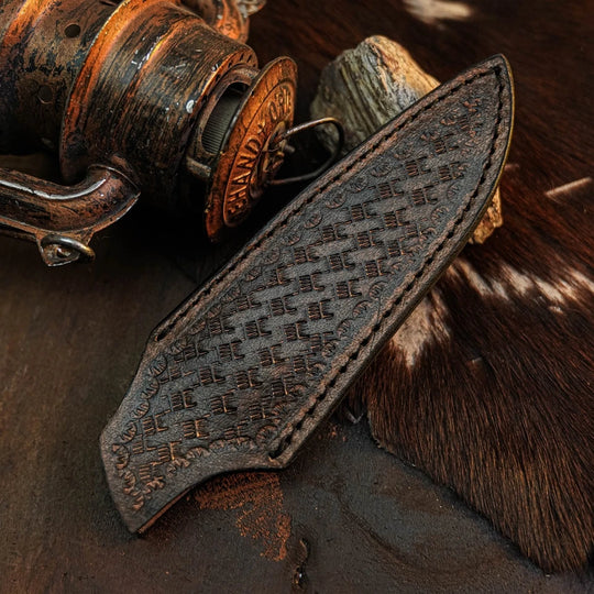 A dark brown leather knife sheath with an embossed geometric pattern, displayed diagonally on a rustic dark surface next to a vintage wooden reel and cowhide.