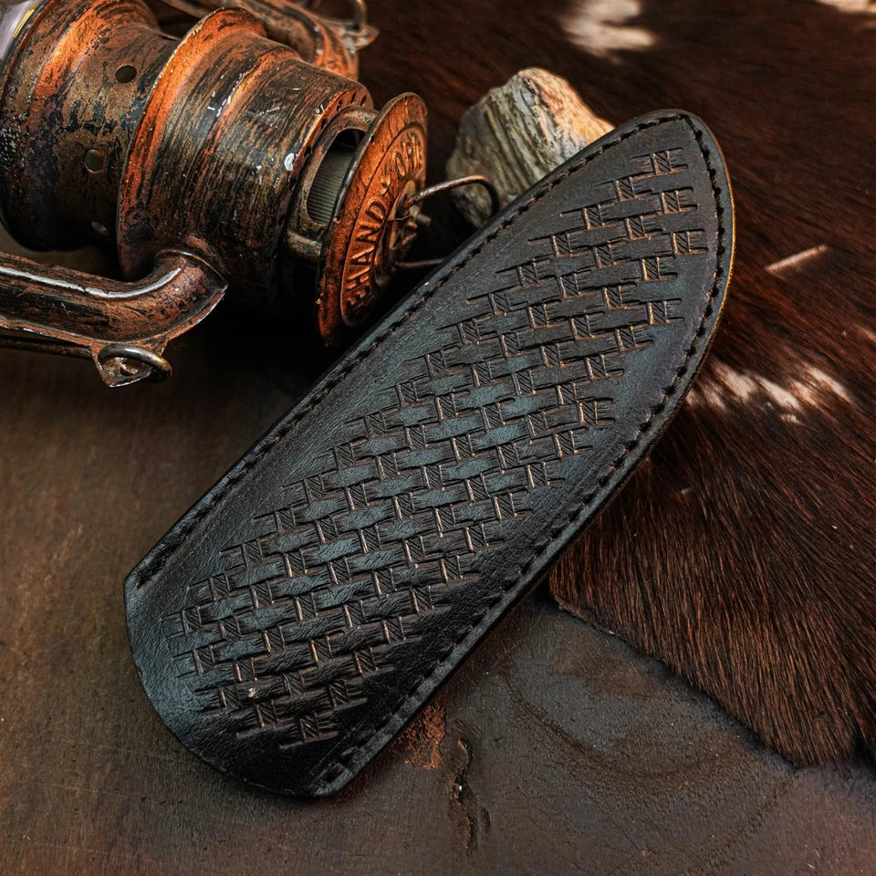 A black leather sheath with an embossed basketweave pattern, shown on a rustic dark wooden surface next to a vintage fishing reel and a piece of brown fur.