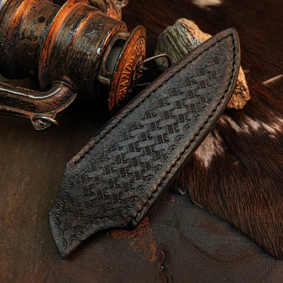 A dark brown leather knife sheath with an embossed basketweave pattern and stitched edges, displayed on a rustic dark surface next to an antique fishing reel and cowhide.