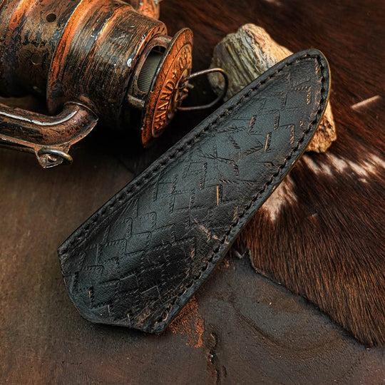 A close-up of a black leather sheath with a textured, embossed pattern and visible stitching, resting on a rustic dark surface next to a vintage copper-toned fishing reel and a brown-and-white hide.