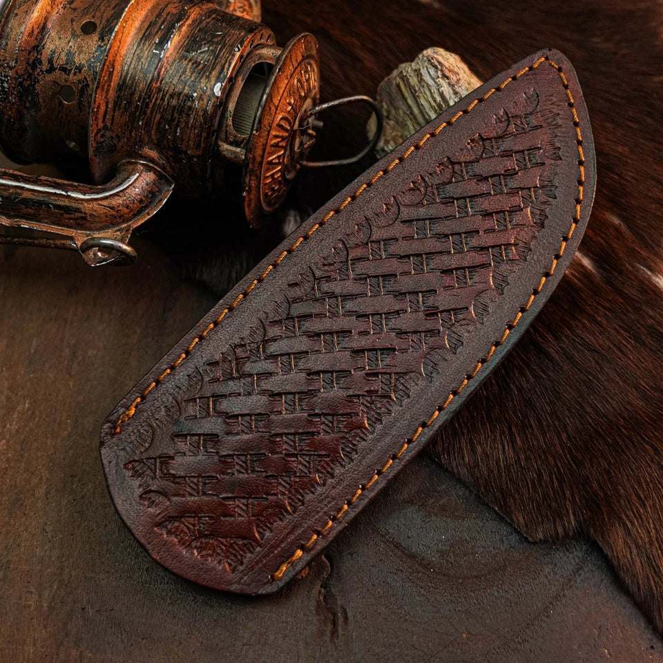 A dark brown leather sheath with an embossed basketweave pattern and orange stitching, displayed diagonally on a rustic background with a vintage fishing reel and fur.