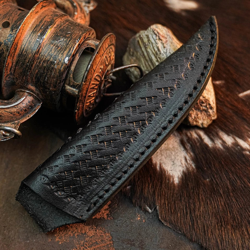 A close-up of a black leather knife sheath with a textured basketweave pattern and visible contrast stitching, resting on a rustic surface with a vintage lantern and cowhide.