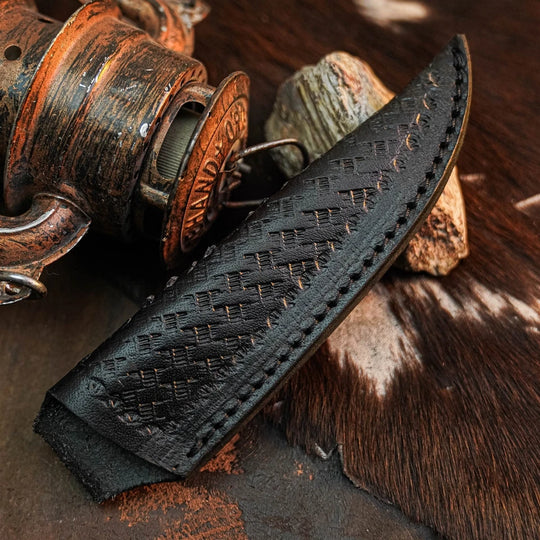A close-up of a black leather knife sheath with a textured basketweave pattern and visible contrast stitching, resting on a rustic surface with a vintage lantern and cowhide.