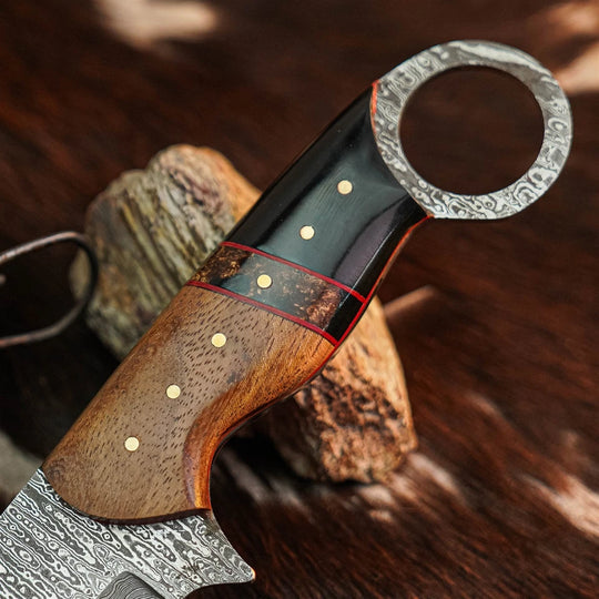 A close-up of a custom Damascus steel knife with a patterned handle, resting on rustic wood blocks against a dark wood background.