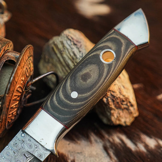 A close-up of a custom knife with a patterned Damascus steel blade and a carved handle featuring a dark wood grain swirl design, resting on rustic rocks.