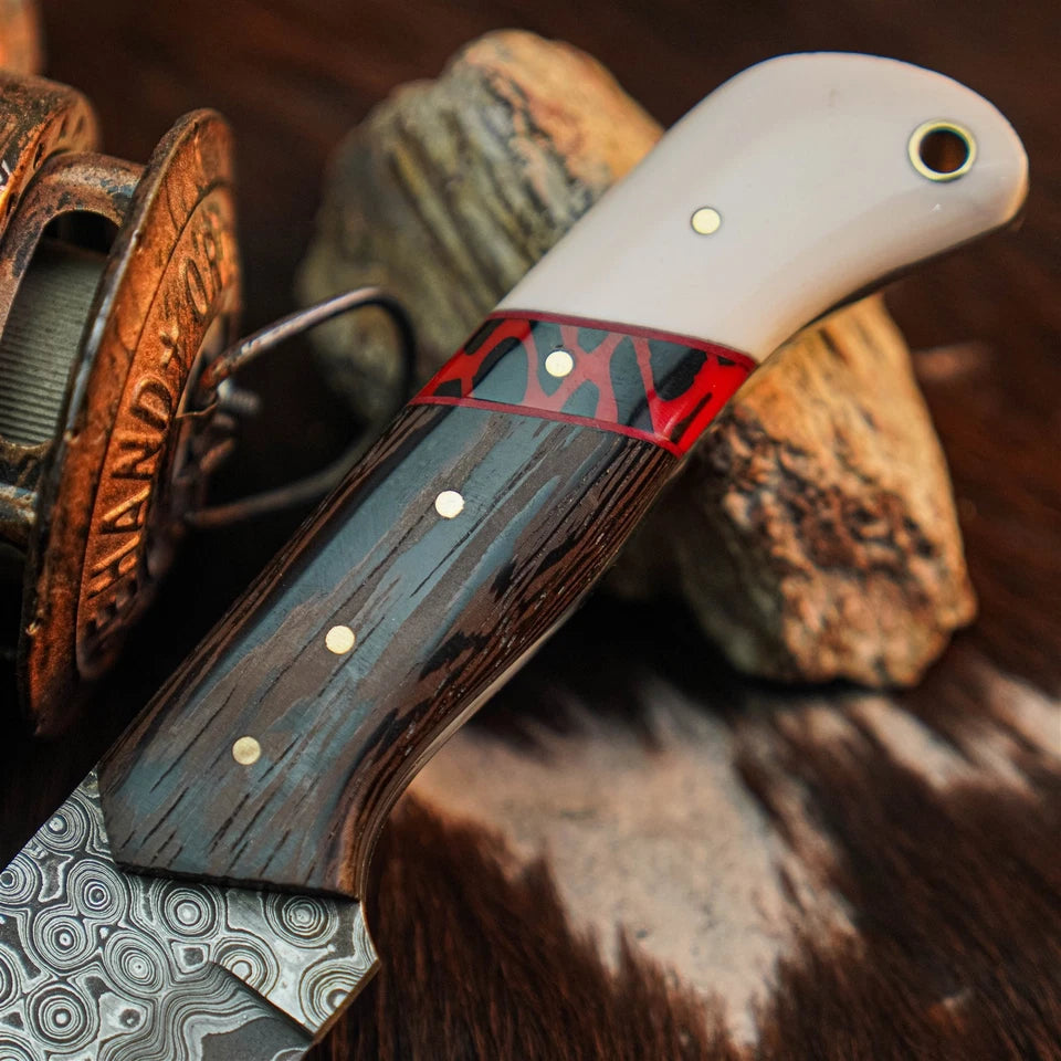 A close-up of a handcrafted knife with a patterned steel blade, dark wood handle, red inlay, and white pommel, resting on rocks with a spool in the background.