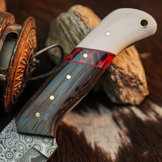 A close-up of a handcrafted knife with a patterned steel blade, dark wood handle, red inlay, and white pommel, resting on rocks with a spool in the background.