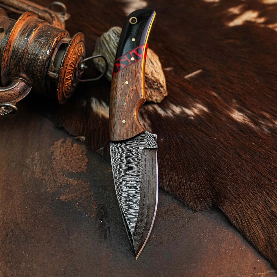 A fixed-blade knife with a patterned Damascus-style steel blade, a brown wood handle with brass pins, and a black end cap with red inlay, resting on a brown fur surface next to a rustic metal object.