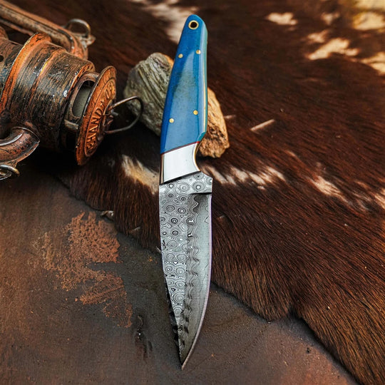 A fixed-blade knife with a patterned steel blade and a blue handle with brass pins, resting on a brown and white cowhide rug next to a rustic wooden object.