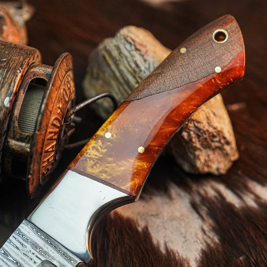 A close-up of a handcrafted knife with a polished reddish-brown and amber wood handle, brass pins, and a satin-finished steel blade, resting on rustic wood and fur.