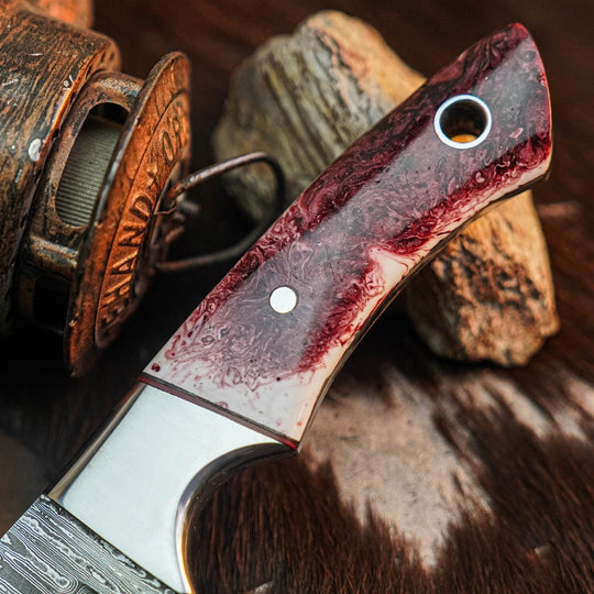 A close-up of a custom knife with a dark red and black marbled handle, resting on a textured hide with rustic wooden props and a brass object in the background.