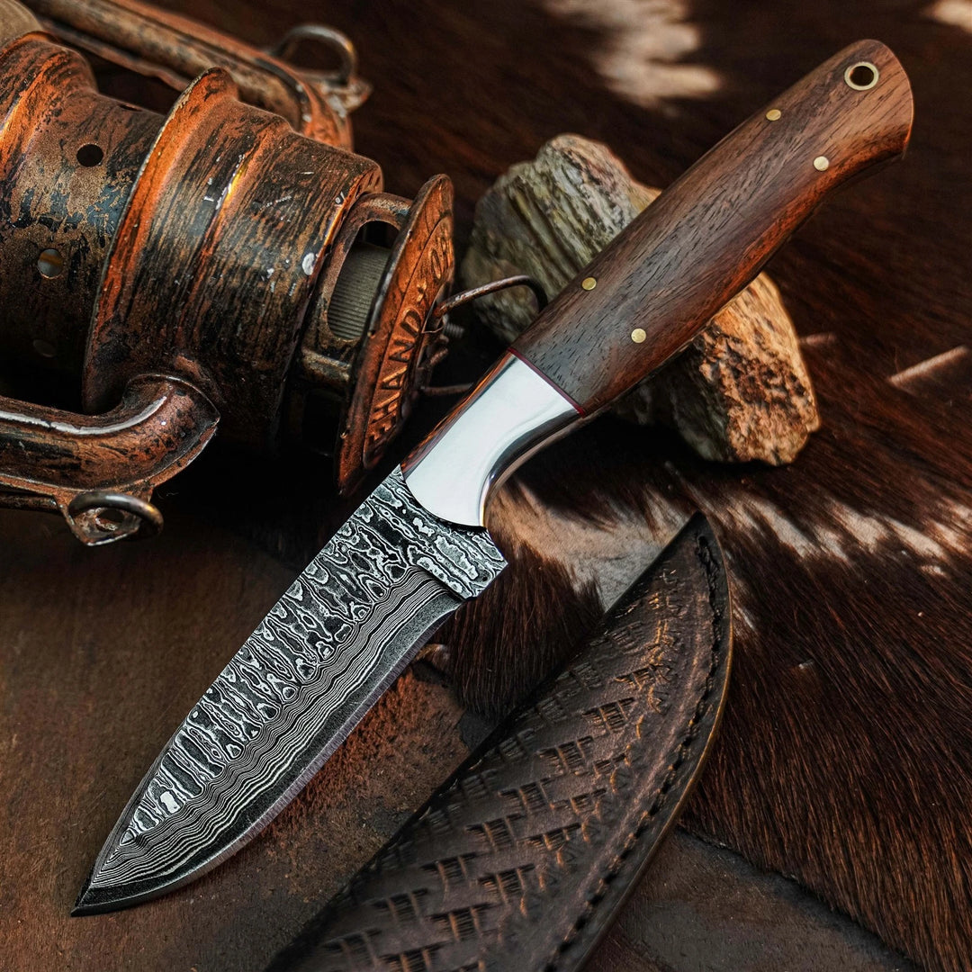 A fixed-blade knife with a patterned Damascus-style steel blade and a dark brown wooden handle, displayed diagonally on a rustic brown hide with a leather sheath and vintage fishing reel in the background.