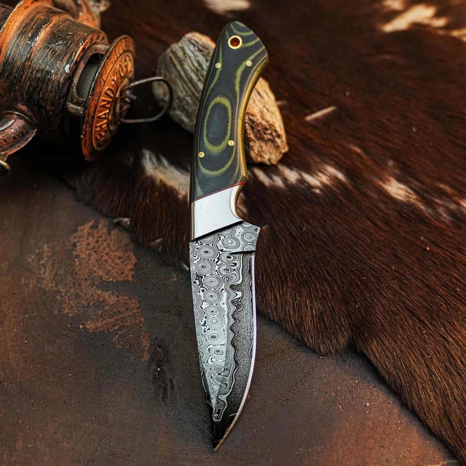 A fixed-blade knife with a patterned steel blade and a dark green handle with gold accents, displayed diagonally on a brown textured surface with a rustic wooden object in the background.