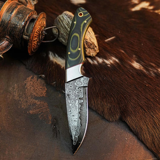 A fixed-blade knife with a patterned steel blade and a dark green handle with gold accents, displayed diagonally on a brown textured surface with a rustic wooden object in the background.