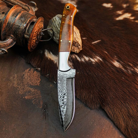 A fixed-blade knife with a patterned Damascus steel blade and a polished brown wood handle, displayed on a dark textured surface next to a rustic wooden object and a cowhide rug.