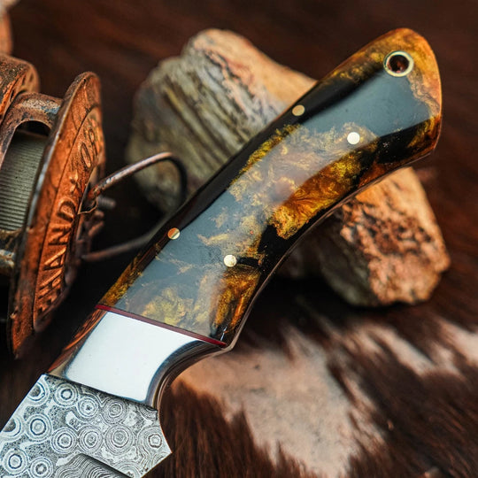 A close-up of a handcrafted knife with a patterned Damascus steel blade and a polished handle made of dark wood with gold and brown inclusions, resting on rustic wood and rocks.