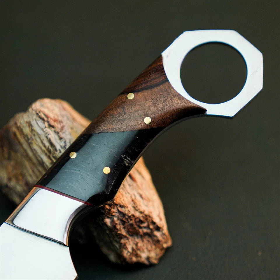 A close-up of a custom knife handle made of dark wood and black inlay with brass pins, resting on a piece of wood against a dark background.