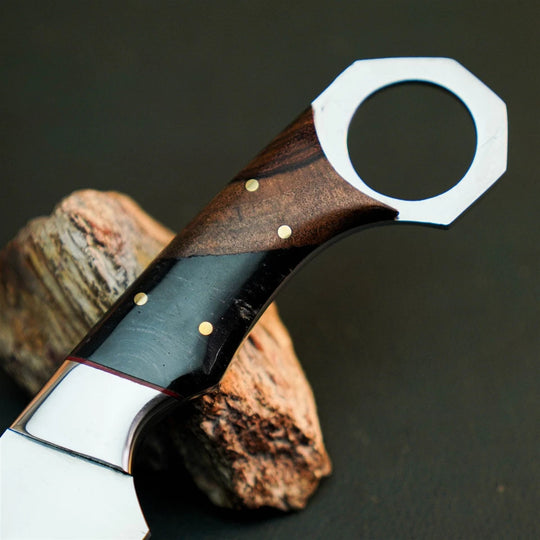 A close-up of a custom knife handle made of dark wood and black inlay with brass pins, resting on a piece of wood against a dark background.