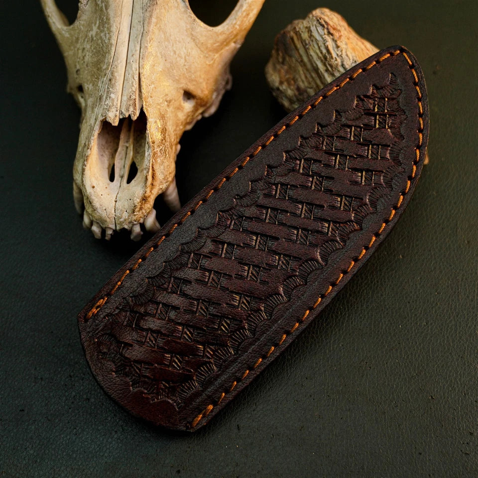 A dark brown leather knife sheath with an embossed basketweave pattern and orange-brown contrast stitching, displayed on a dark textured surface with animal skull props in the background.