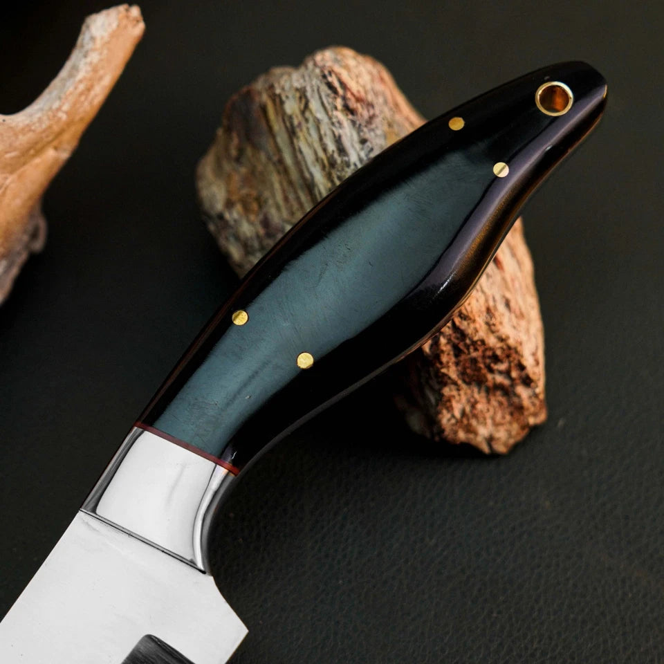 A close-up of a knife with a polished silver blade and a dark handle featuring a blue-green inlay, gold pins, and brass pommel, resting on textured rocks against a dark background.