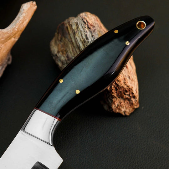 A close-up of a knife with a polished silver blade and a dark handle featuring a blue-green inlay, gold pins, and brass pommel, resting on textured rocks against a dark background.
