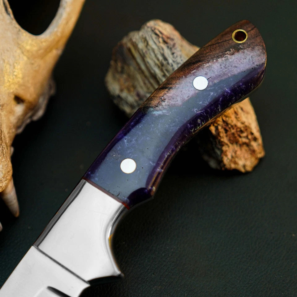 A close-up of a fixed-blade knife with a polished silver blade and a dark purple and brown handle, resting on natural wood pieces against a dark background.