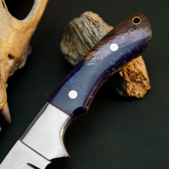 A close-up of a fixed-blade knife with a polished silver blade and a dark purple and brown handle, resting on natural wood pieces against a dark background.